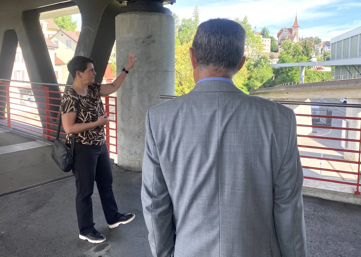 Eine Frau in gemusterter Bluse steht auf einer gedeckten Überführung und zeigt auf den Hintergrund. Ein Mann im hellgrauen Anzug hört ihr zu.